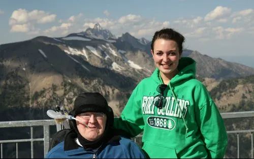 Man in wheelchair with a ventilation device and woman in green sweatshirt pose for a photo in front of a mountain range on a sunny day